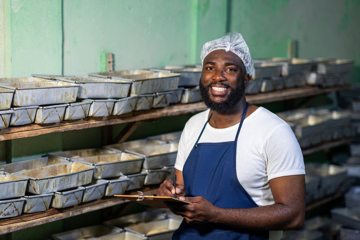 Portrait of smiling local African baker entrepreneur in a hairnet and apron, holding a clipboard in his bakery, successful small business owner SME